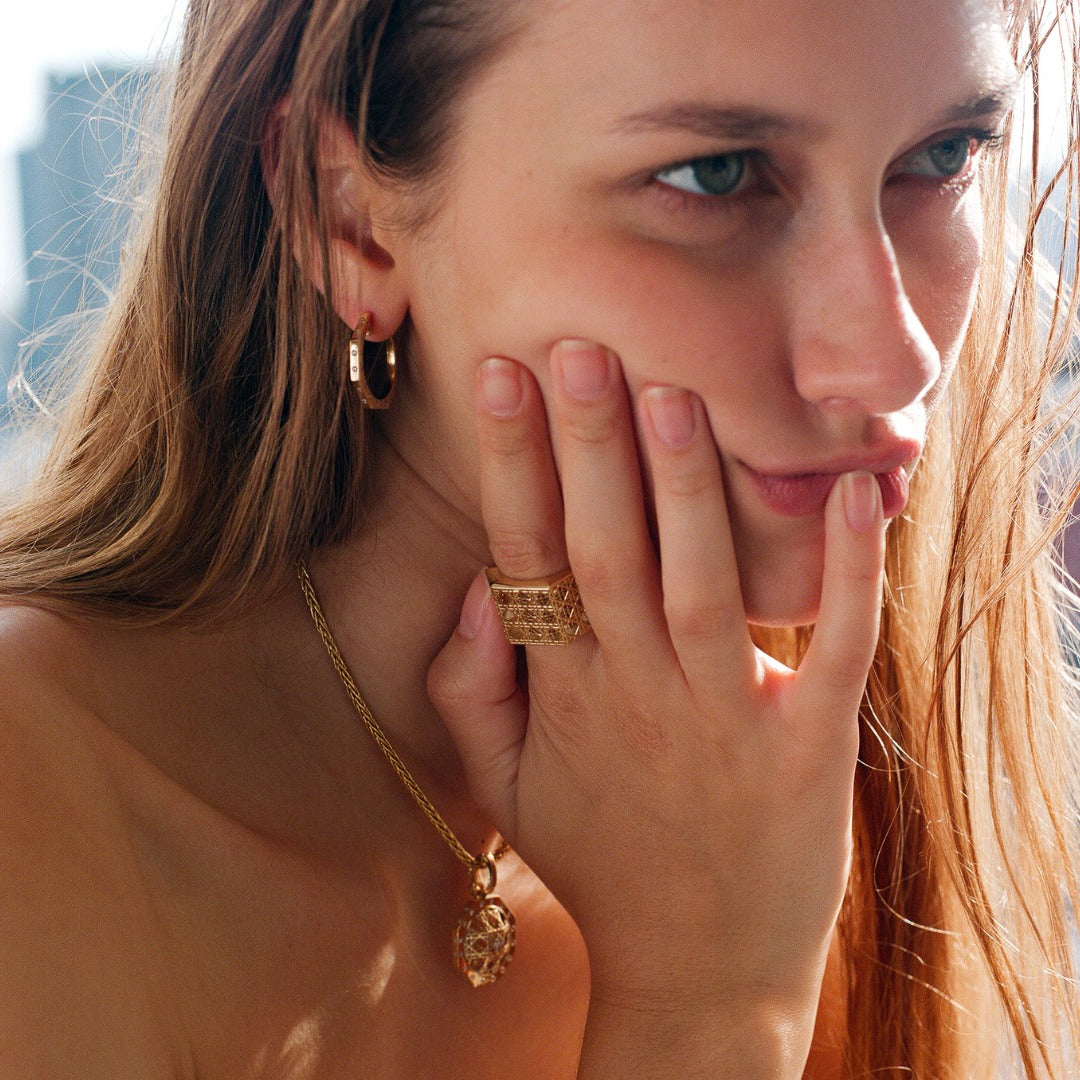 MoAnA LUU fine jewelry worn on model, featuring Ti hoop earrings and Doudou wide ring and pendant in 18K yellow gold inspired by Caribbean fish traps.