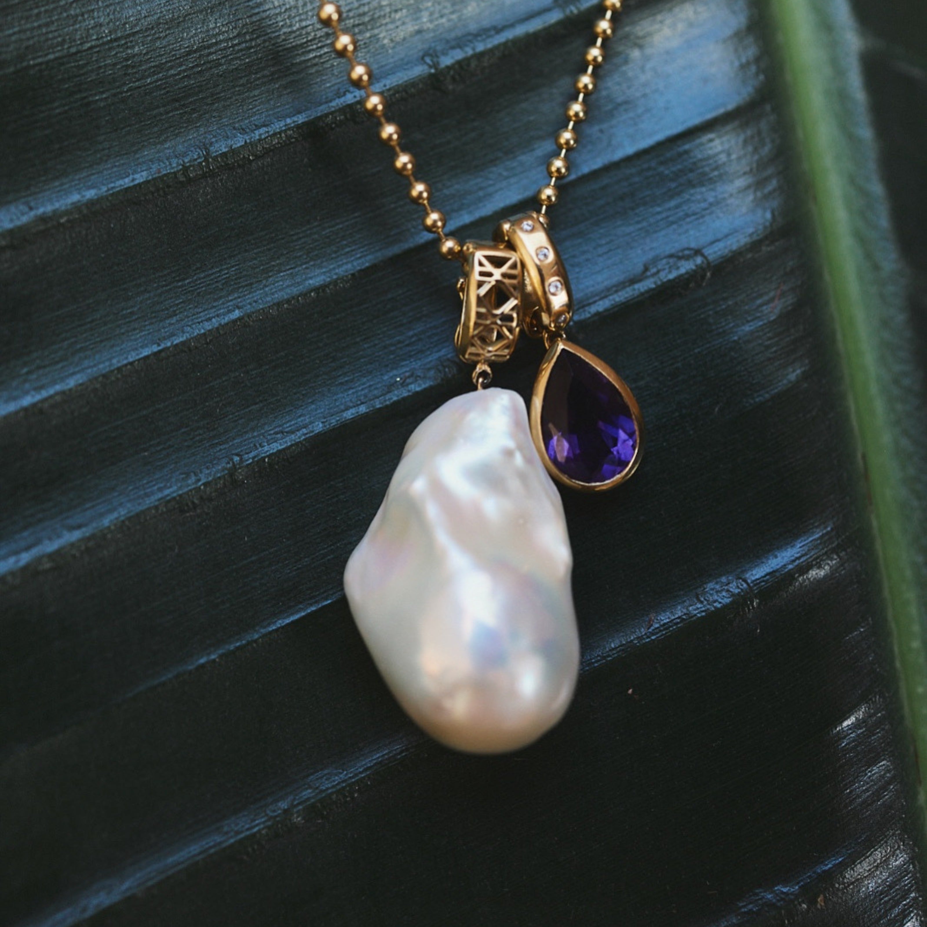 MoAnA LUU Abyss baroque pearl pendant and Dlo amethyst pendant in 18K yellow gold on green leaf. 