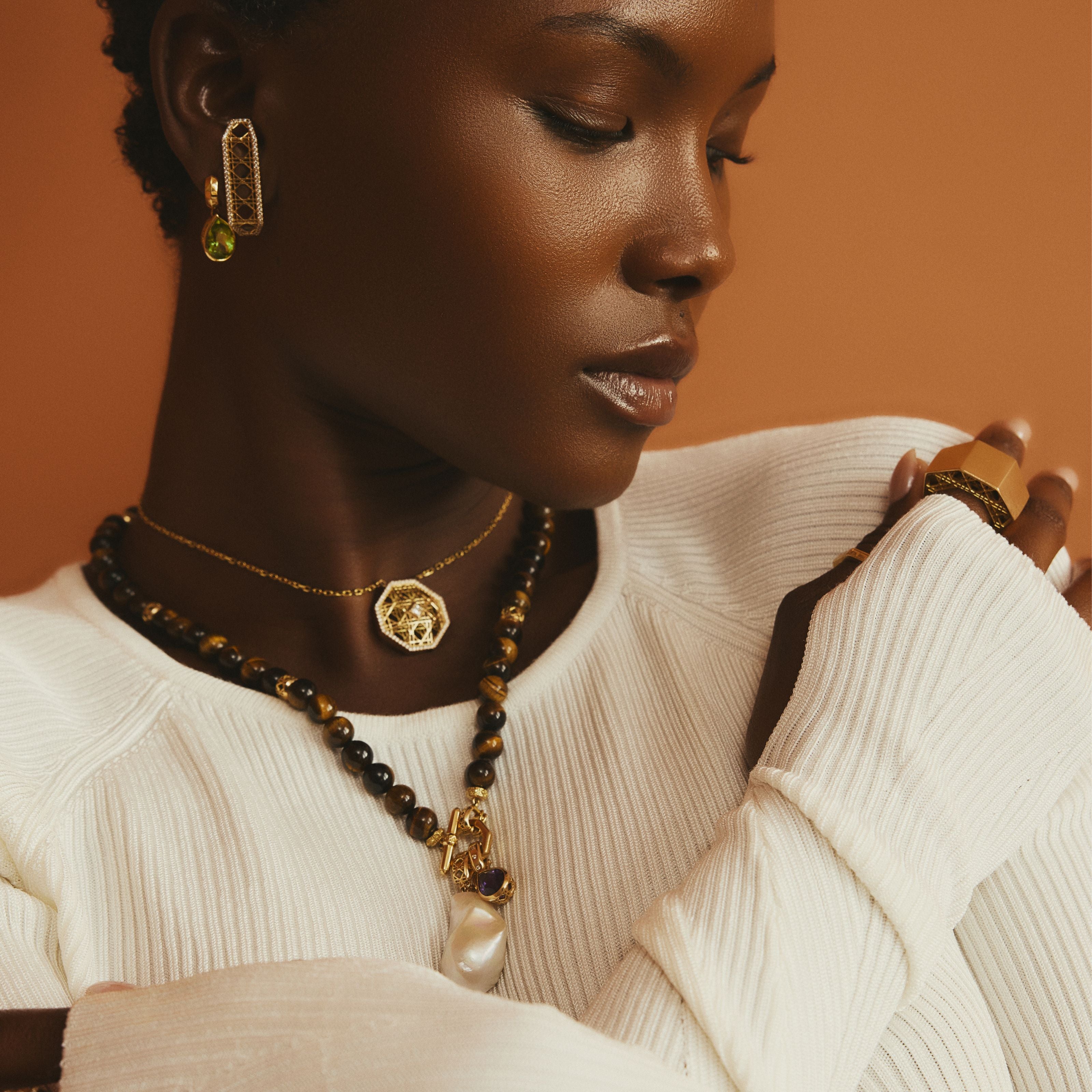 MoAnA LUU fine jewelry in 18K yellow gold worn layered on model, featuring tiger eye Yva bead necklace, Doudou pendant, barroque pearl charm, Dlo and Doudou earrings, and sculptural gold Doudou signet ring inspired by Creole design.