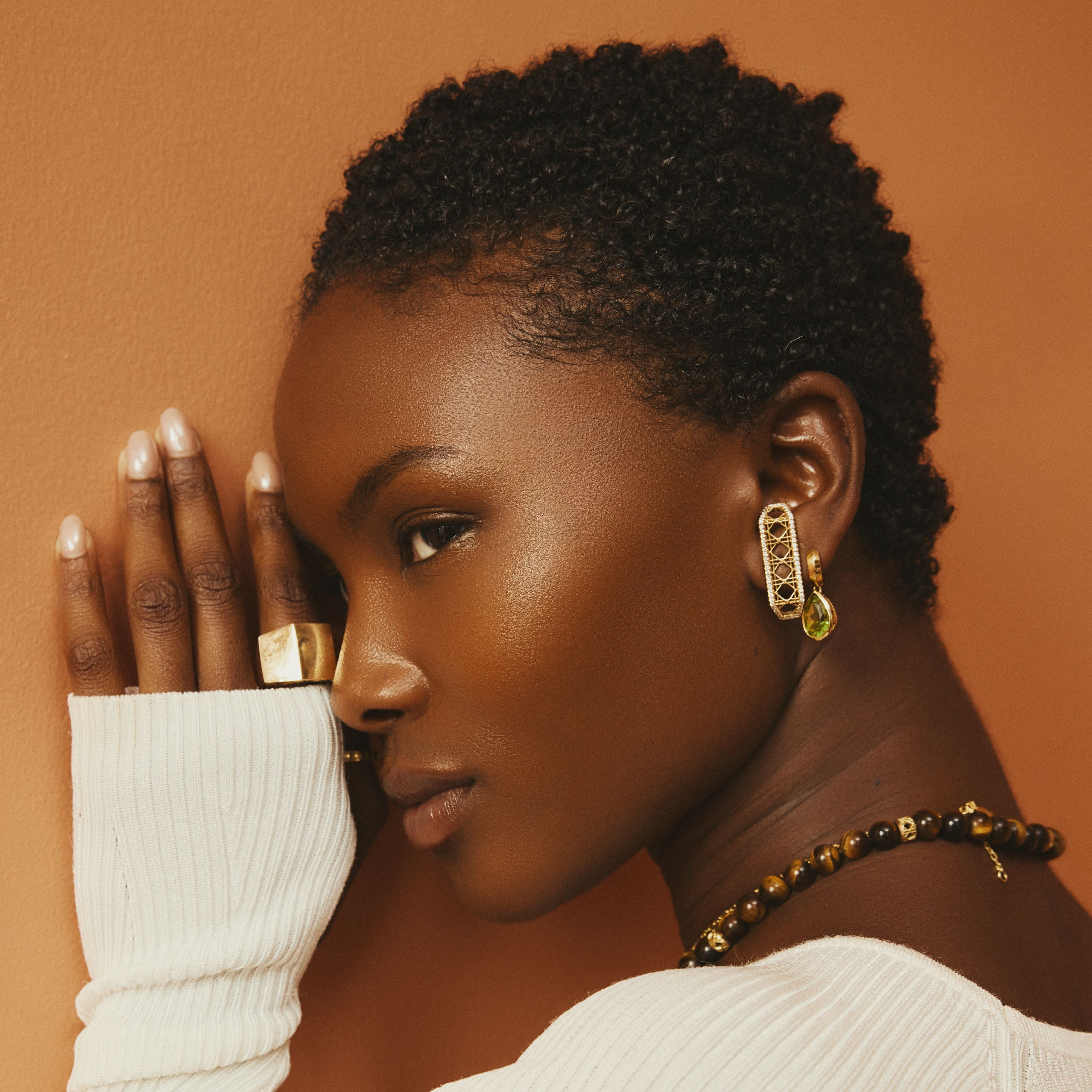 MoAnA LUU fine jewelry in 18K yellow gold worn layered on model, featuring tiger eye Yva bead necklace, Dlo and Doudou earrings, and sculptural gold Doudou signet ring inspired by Creole design.