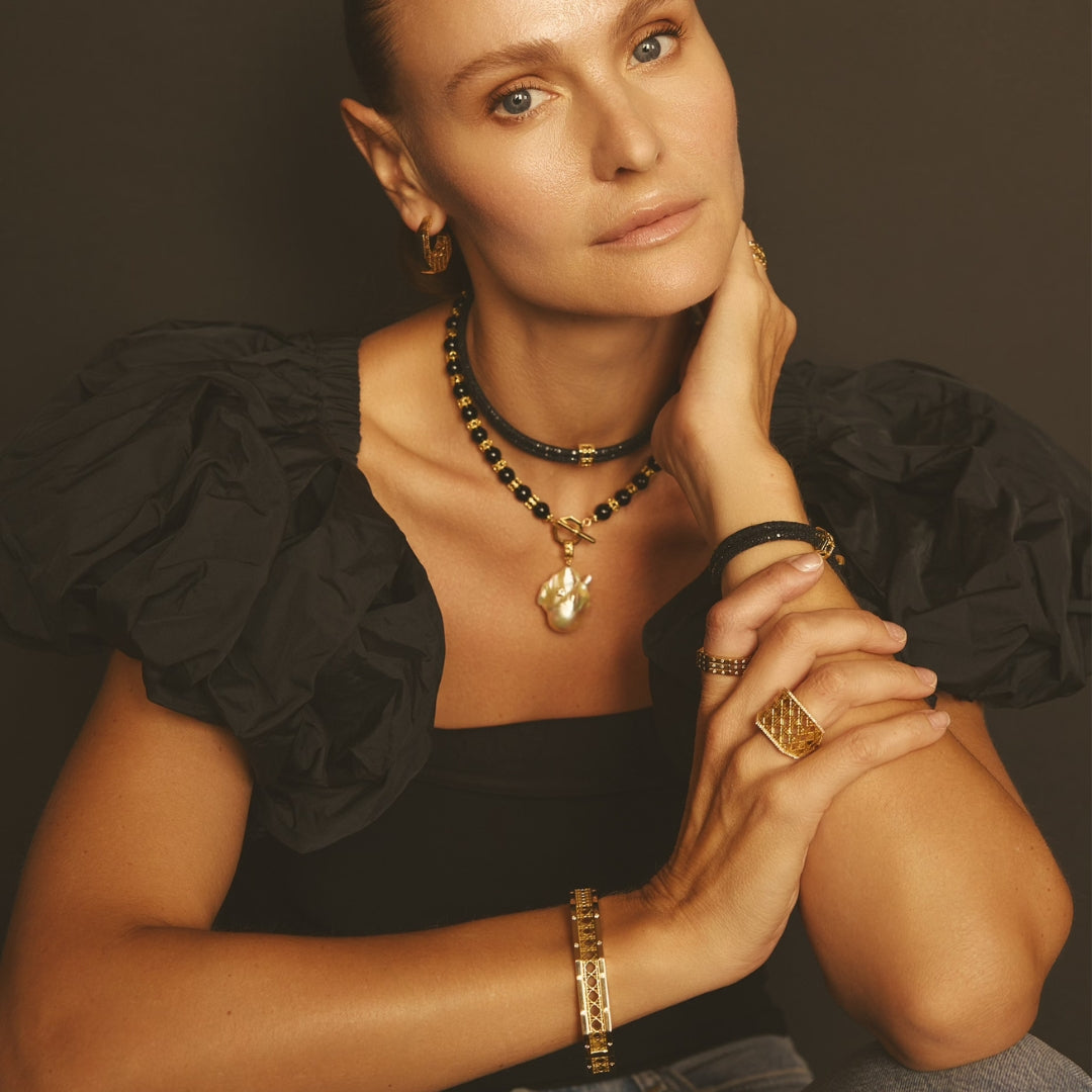 MoAnA LUU fine jewelry in 18K yellow gold worn layered on model, featuring Yva onyx bead necklace, Abyss baroque pearl pendant, Abyss stingray leather choker and bracelet, and sculptural Doudou rings, bracelet, and hoops inspired by Creole design.